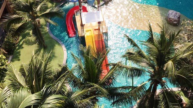 Un espace piscine d’hôtel avec un toboggan aquatique, des palmiers et un bâtiment doté de balcons.