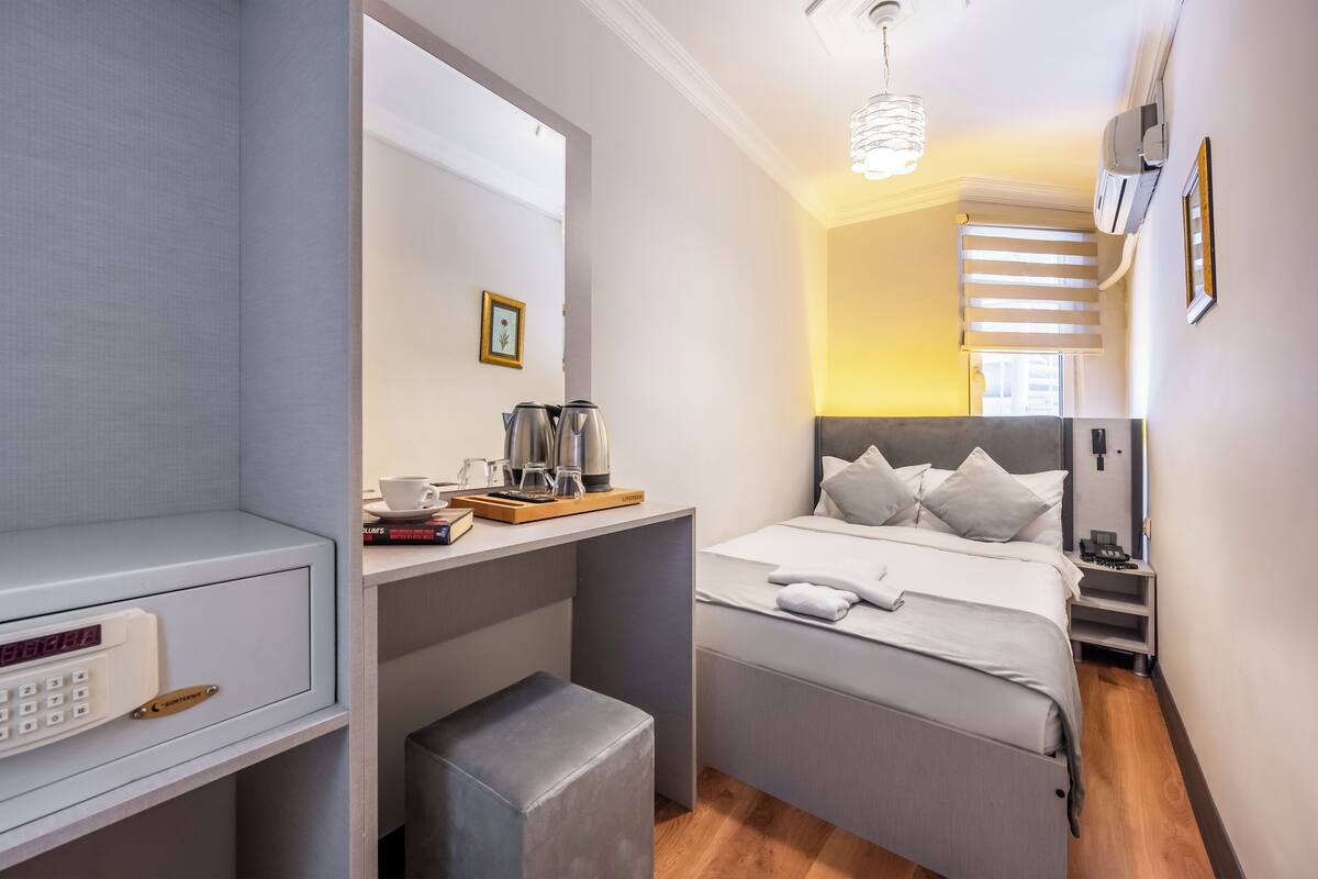 Ein kleines Hotelzimmer mit Bett, Nachttisch und einer kleinen Sitzecke mit einer Kaffeemaschine und Büchern.