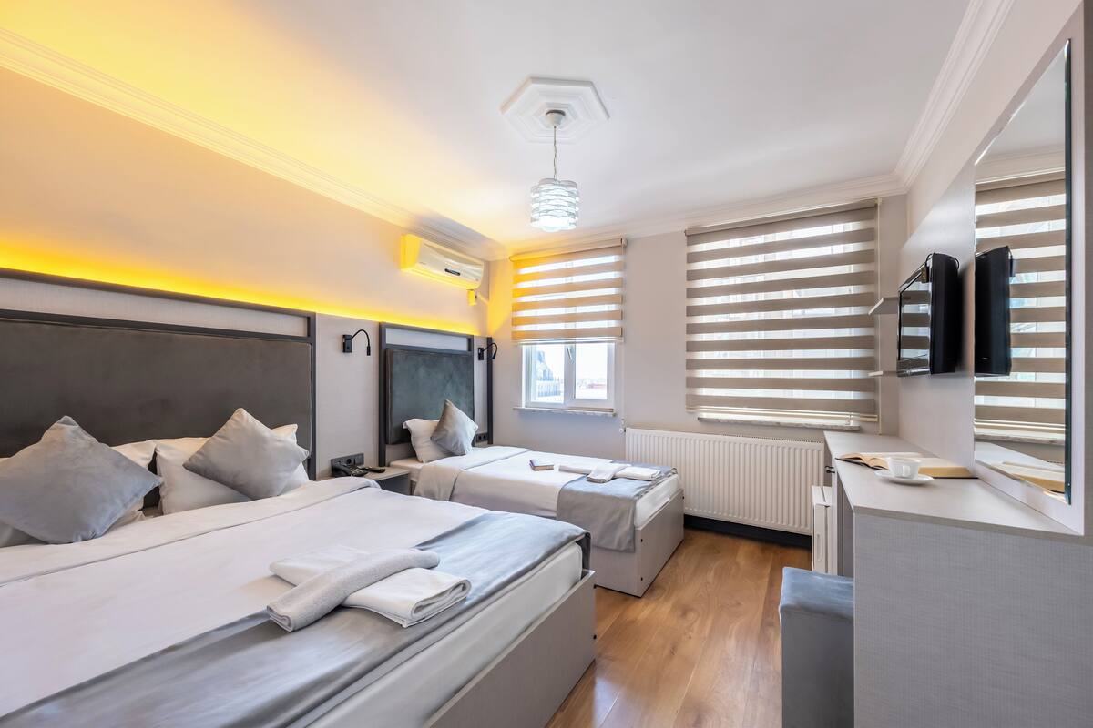 Ein modernes Hotelzimmer mit zwei Betten, einem Schreibtisch, einem Fernseher und großen Fenstern mit Jalousien.