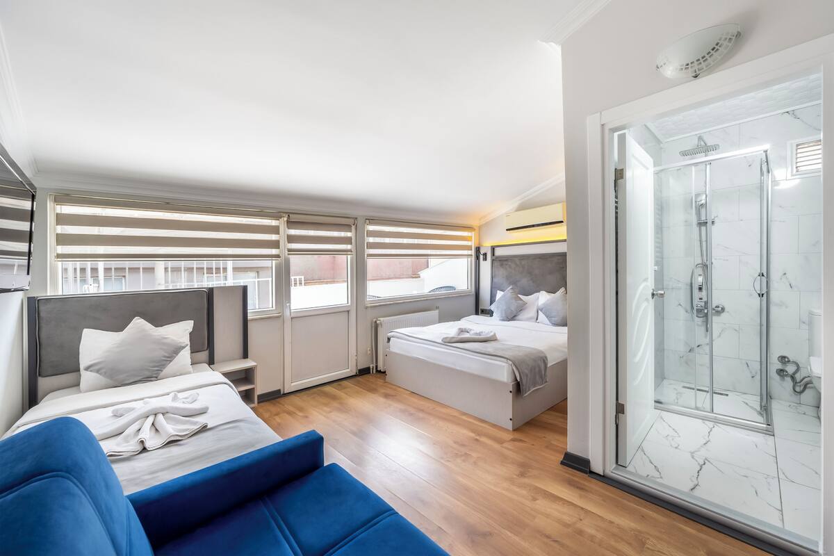 Ein modernes Hotelzimmer mit einem großen Bett, einer separaten Sitzecke, einer Duschkabine aus Glas und Blick auf die Stadt durch die Fenster.