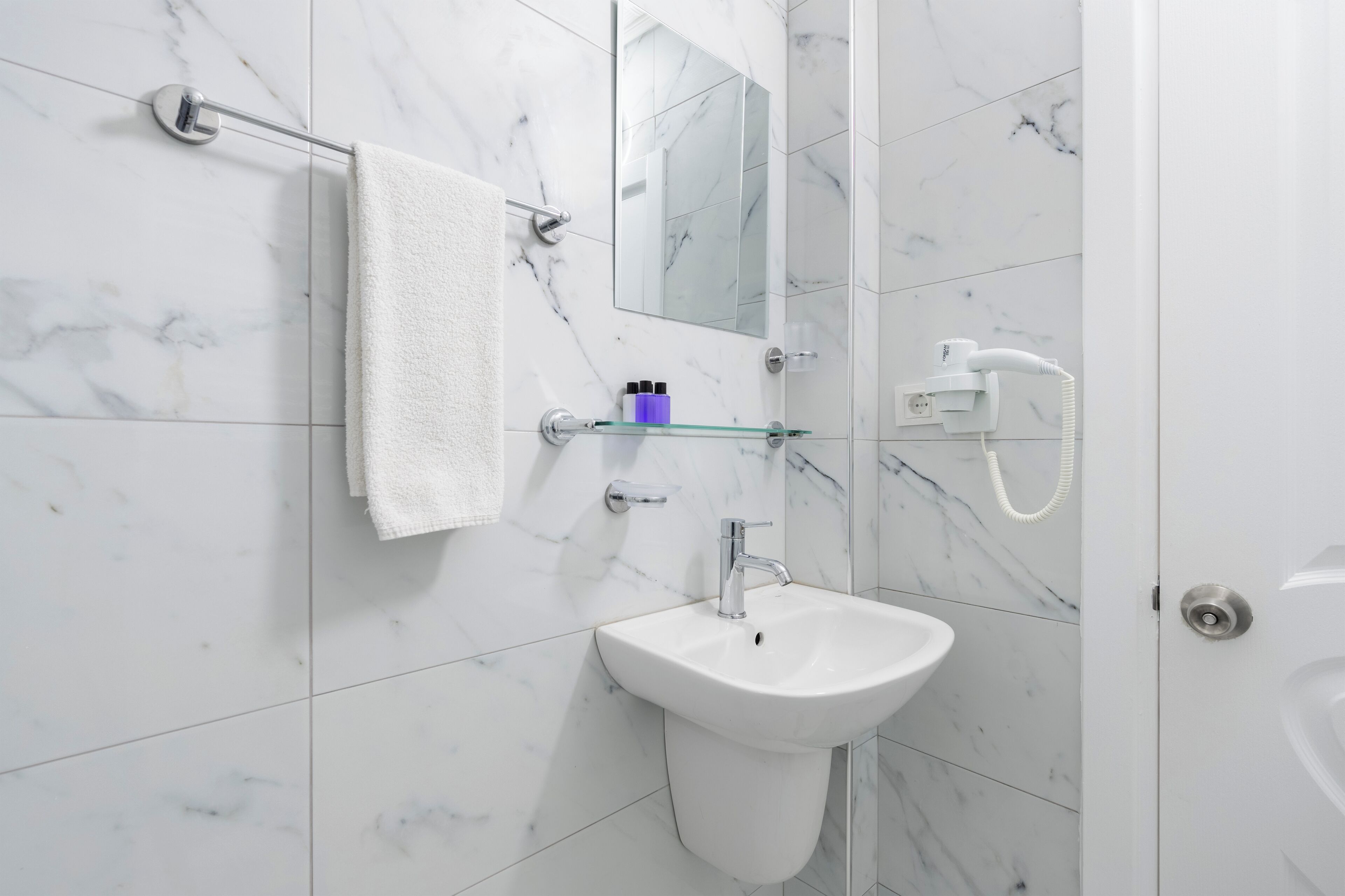 Une salle de bain avec un lavabo en marbre blanc, une serviette suspendue à un porte-serviettes, une étagère en verre contenant des articles de toilette et un miroir au-dessus du lavabo.