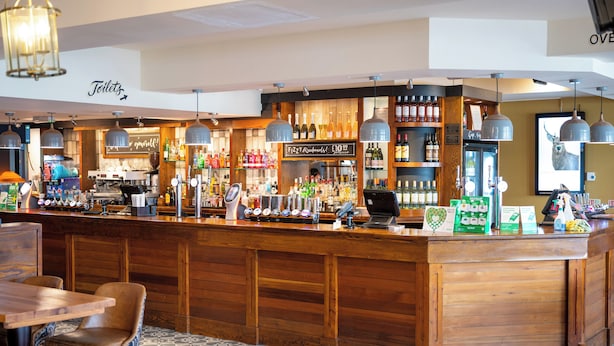 Zona de bar con mostrador de madera, asientos y diferentes botellas y vasos.