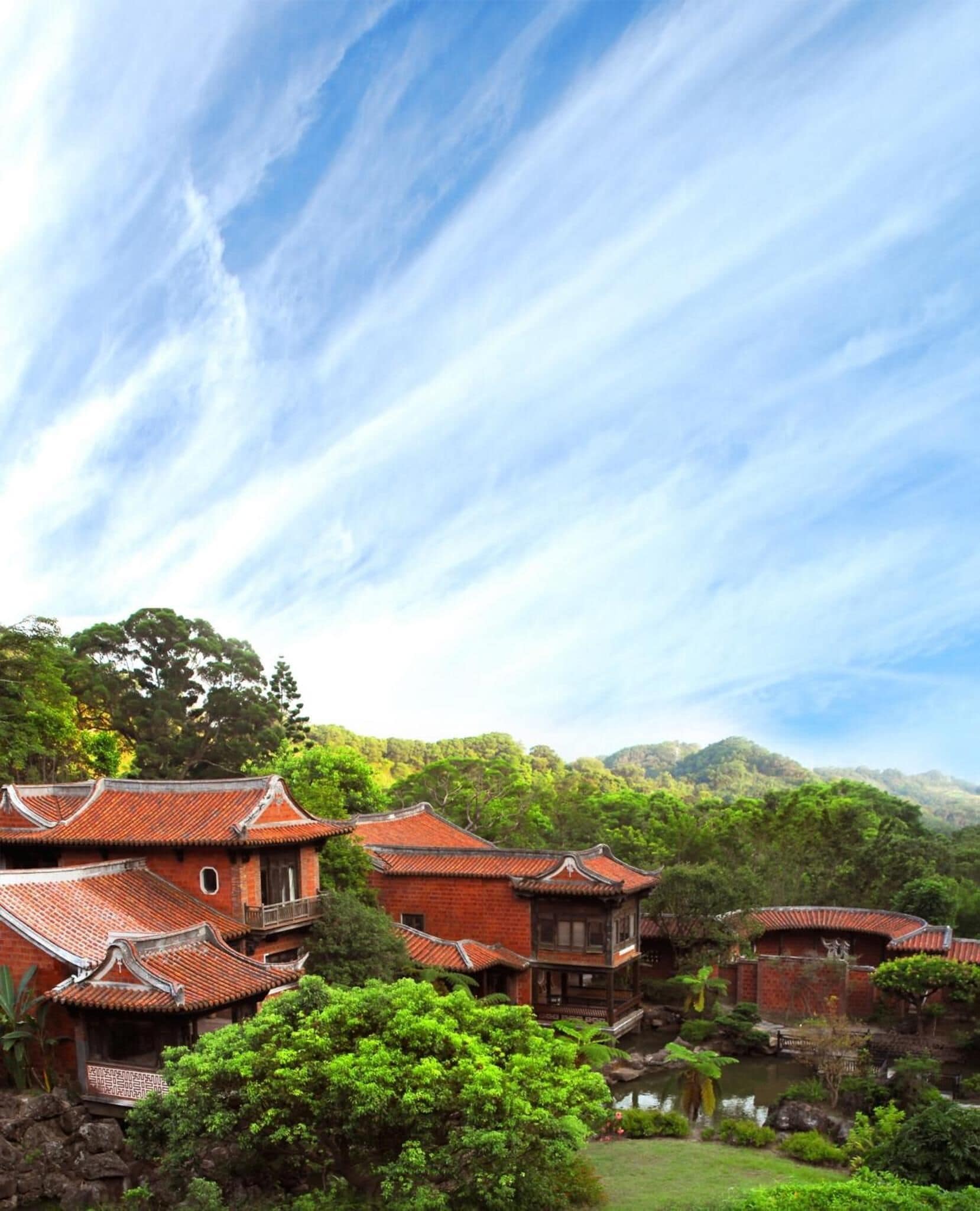 A traditional Chinese courtyard with red-tiled roofs, surrounded by lush greenery and hills.