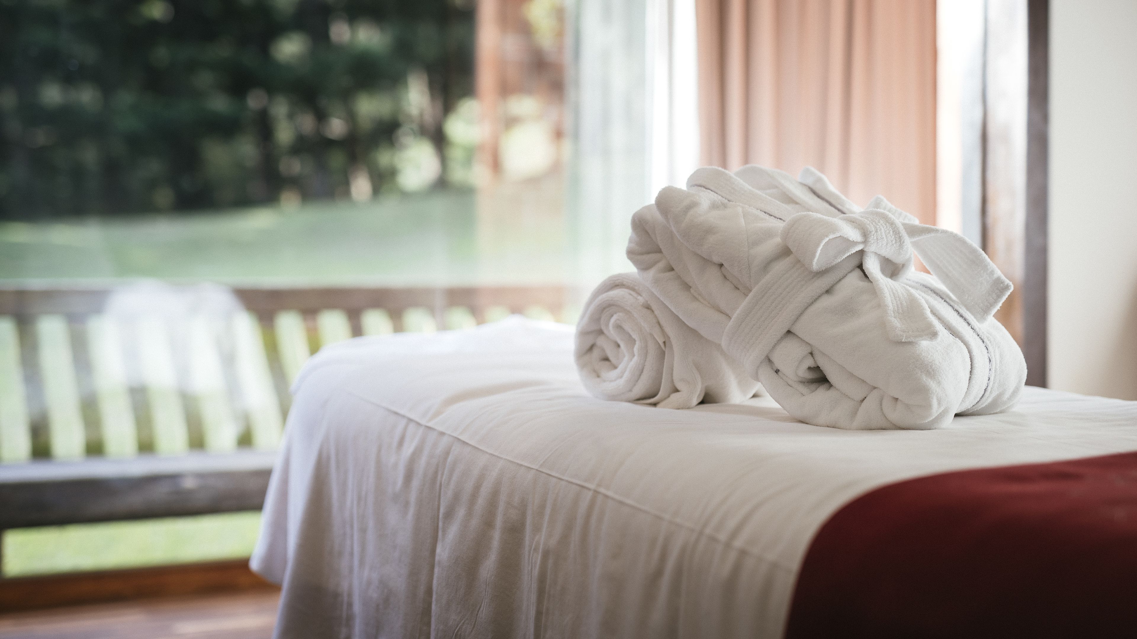 A neatly made bed with a white comforter and a red blanket, a stack of white bathrobes, and a view of a wooden deck and greenery through a window.