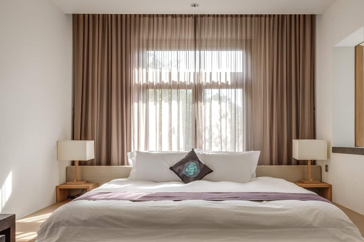 A hotel room with a large bed, two bedside tables with lamps, a window with curtains, and a mirror on the wall.