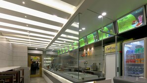 A modern dessert counter with illuminated menu boards, a self-service beverage dispenser, and a clear view into the kitchen area. - Hotel Sifat International (Surat)