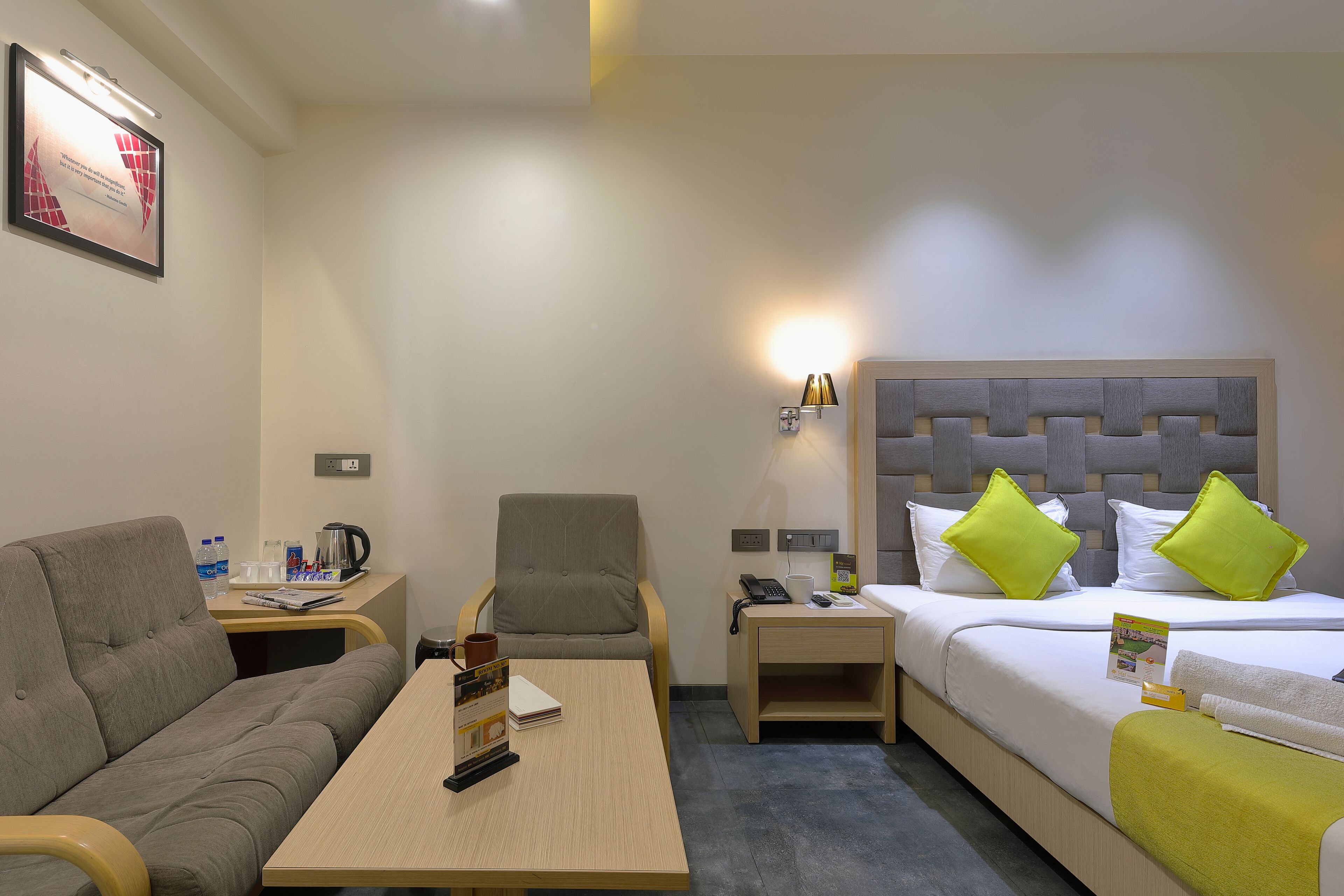 Habitación de hotel con sofá, una pequeña mesa con un control remoto, cama con almohadas, mesita de noche con teléfono y lámpara, y un cuadro colgado en la pared.