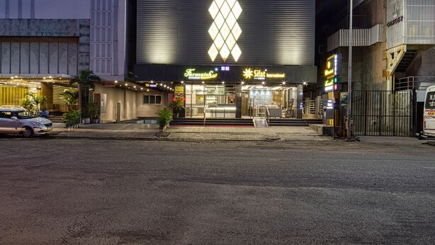 A modern building with a prominent vertical light pattern on the facade, a parking lot in front, and a street with a few cars. - Hotel Sifat International (Surat)