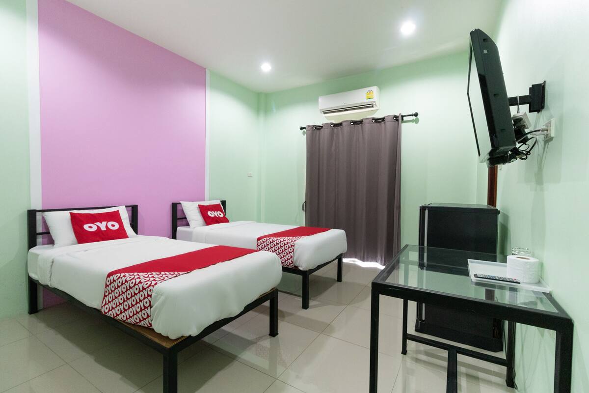 A hotel room with two beds, a desk, a TV, and an air conditioning unit.