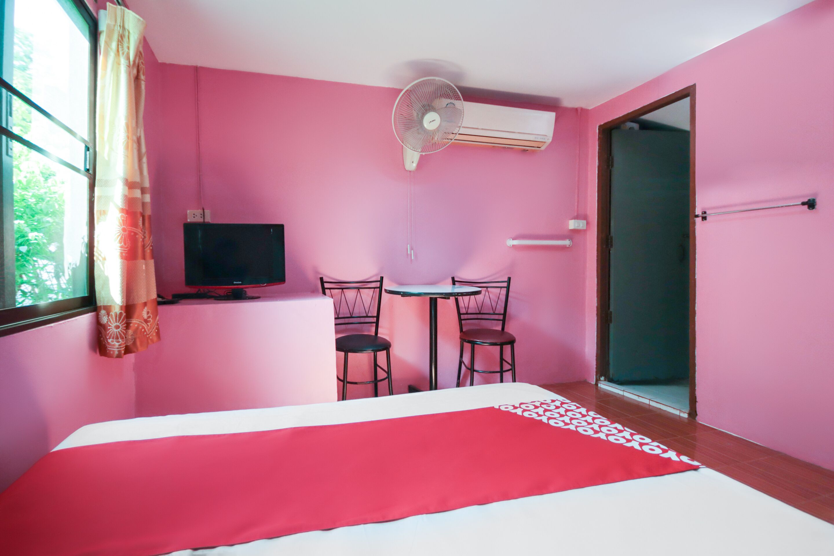 A room with pink walls, a bed with a red and white bedspread, a TV on a stand, two chairs around a small table, and an air conditioning unit.