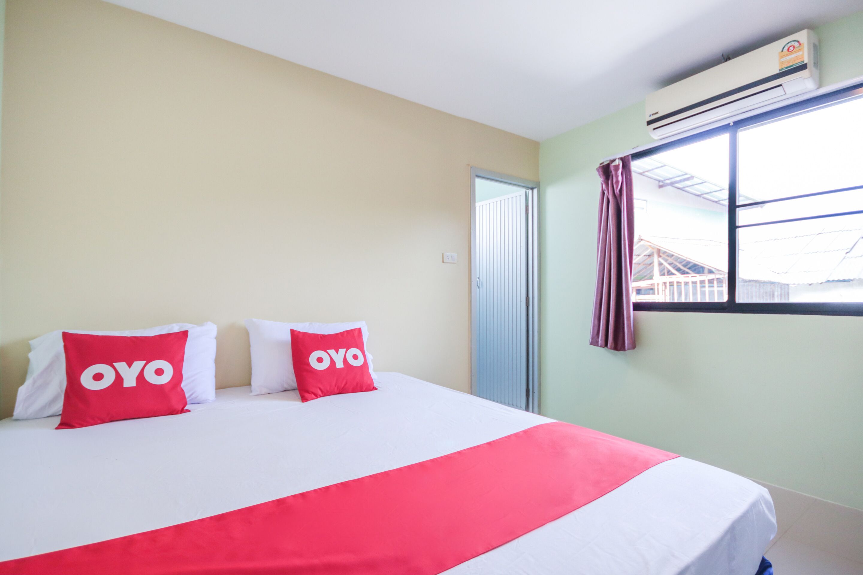 A hotel room with a bed, two pillows with the logo "OYO", a window with a curtain, and an air conditioning unit.