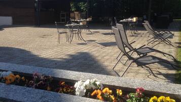 Un'area patio all'aperto con tavoli e sedie, una zona salotto coperta e un'aiuola con diversi fiori.