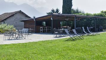A covered outdoor seating area with tables and chairs, a building with a sloped roof, and a grassy lawn.
