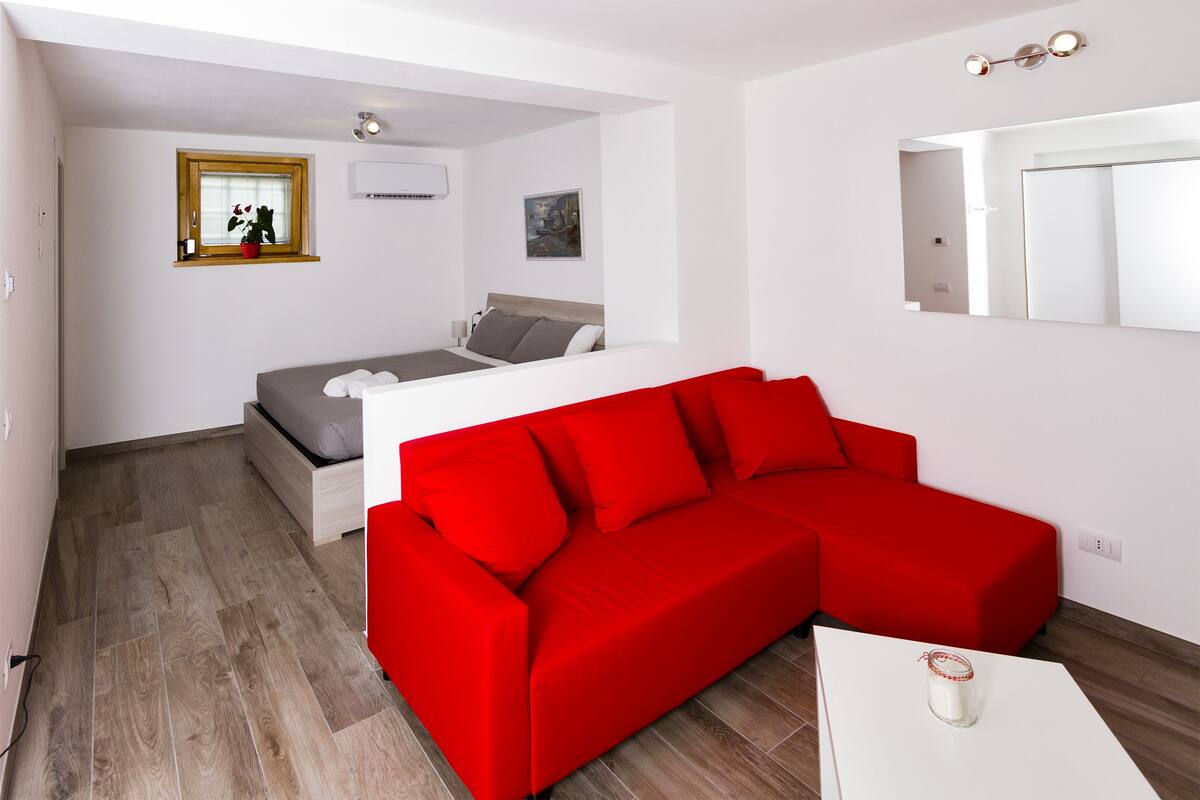 Ein modernes, kompaktes Zimmer mit einem roten Sofa, einem Bett mit grauer Bettwäsche, einem kleinen Tisch mit einem Glas und einem Fenster mit einer Pflanze.