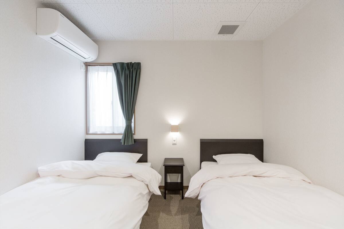 A hotel room with two single beds, a small bedside table, a wall-mounted air conditioner, a window with curtains, and a wall-mounted light fixture.