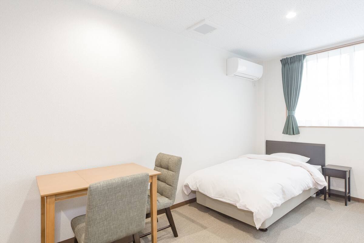 A hotel room with a bed, a desk, a chair, a nightstand, and an air conditioning unit.