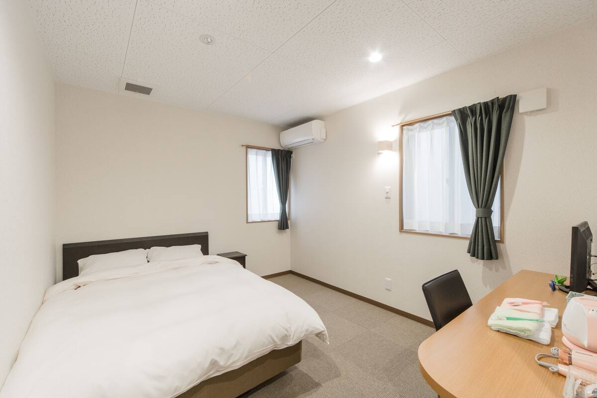 A hotel room with a bed, a desk with a computer, and two windows with curtains.