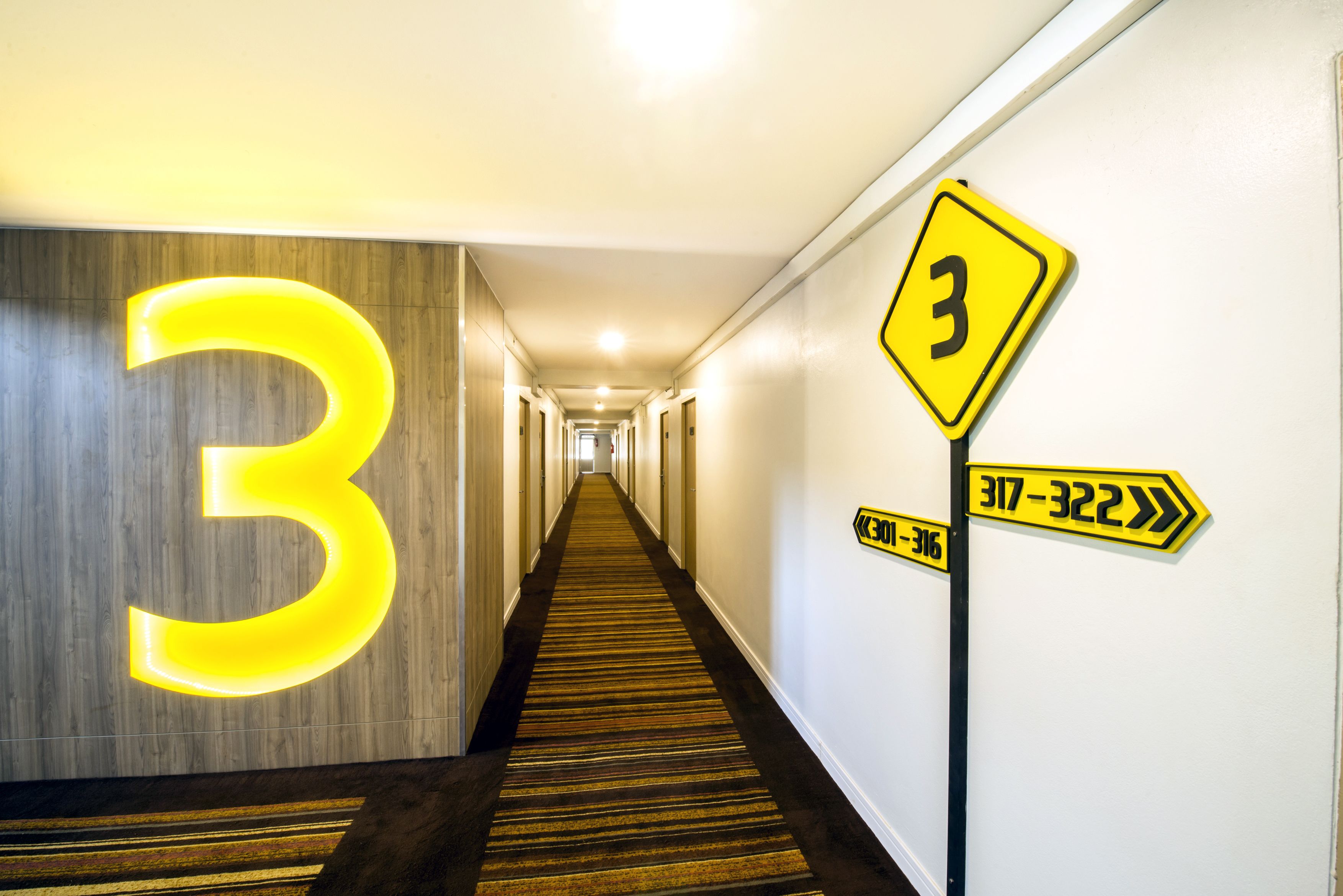 A hotel hallway with a large illuminated number '3' on the wall, directional signs, and a carpeted floor.