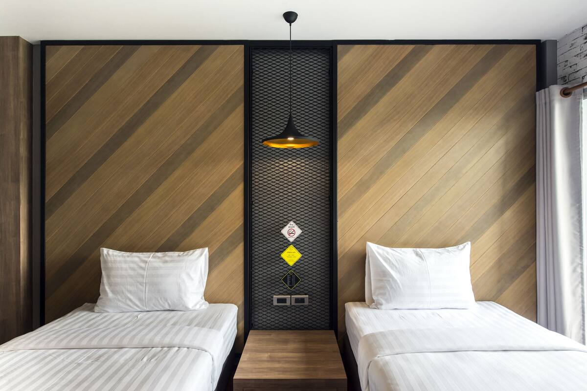 A hotel room with two beds, a central wall panel with a hanging light, and a wooden headboard.