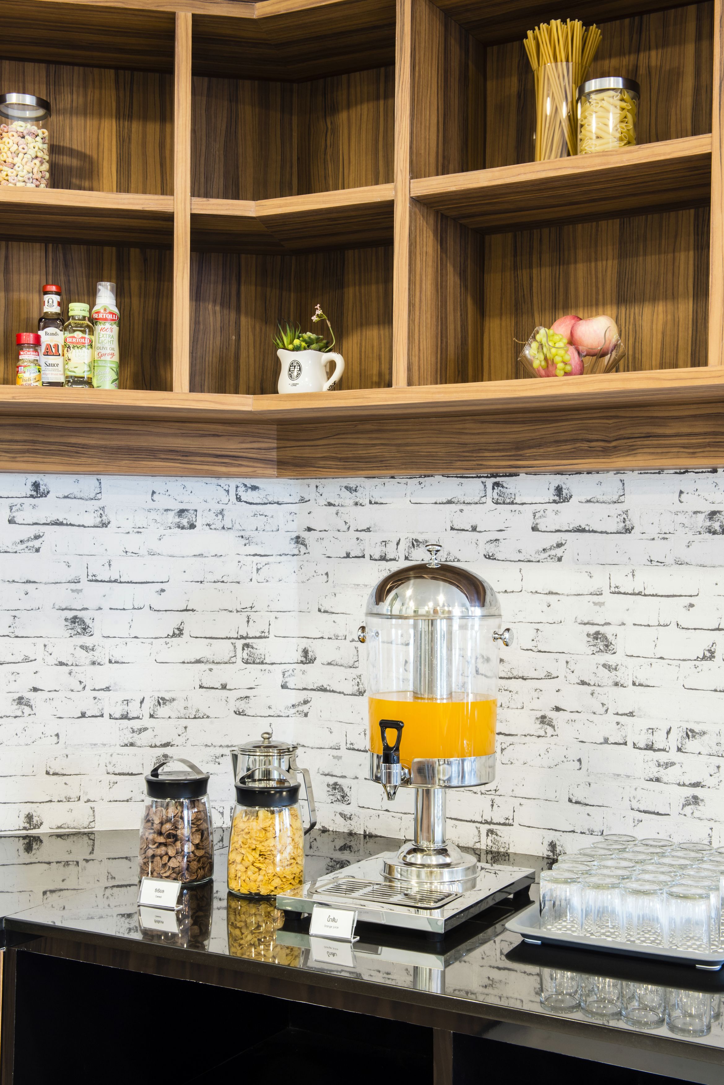 A buffet setup with a coffee dispenser, labeled containers of food, and a brick-patterned wall.
