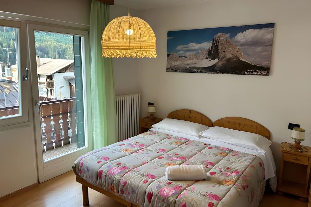 Un dormitorio con cama, lámpara colgante, radiador, mesita de noche y ventana con vistas a edificios y árboles.