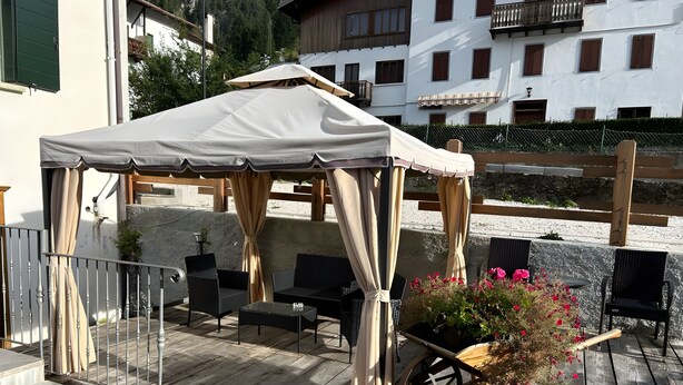 A covered outdoor seating area with a table and chairs, a potted plant, and a wooden cart. - Meublè Bar Giustina (Auronzo di Cadore)