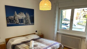 A bedroom with a large bed, a nightstand, a lamp, a radiator, and a window with a view of buildings and cars. - Meublè Bar Giustina (Auronzo di Cadore)