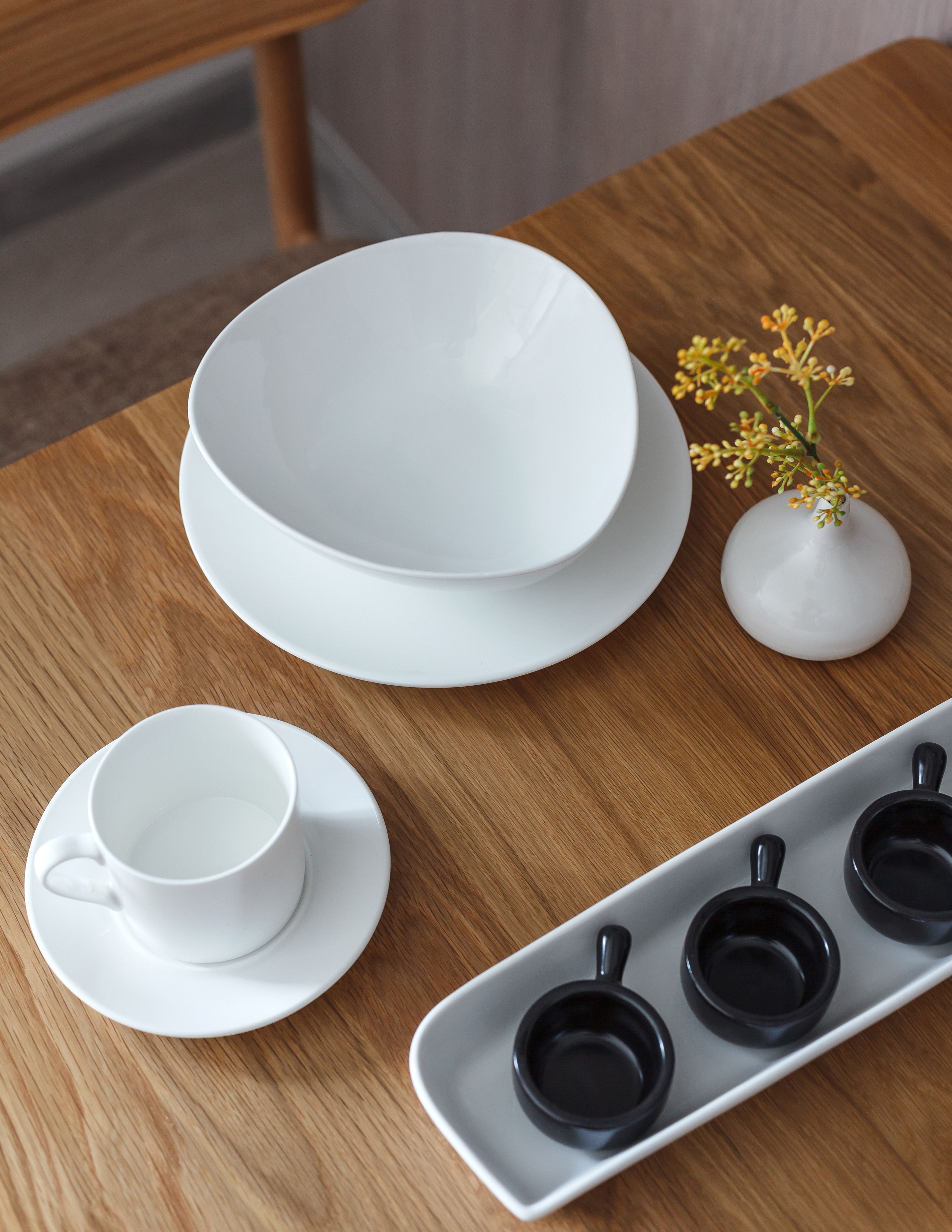 A wooden table with a white bowl, a white cup and saucer, a small vase with yellow flowers, and a tray with black cups.
