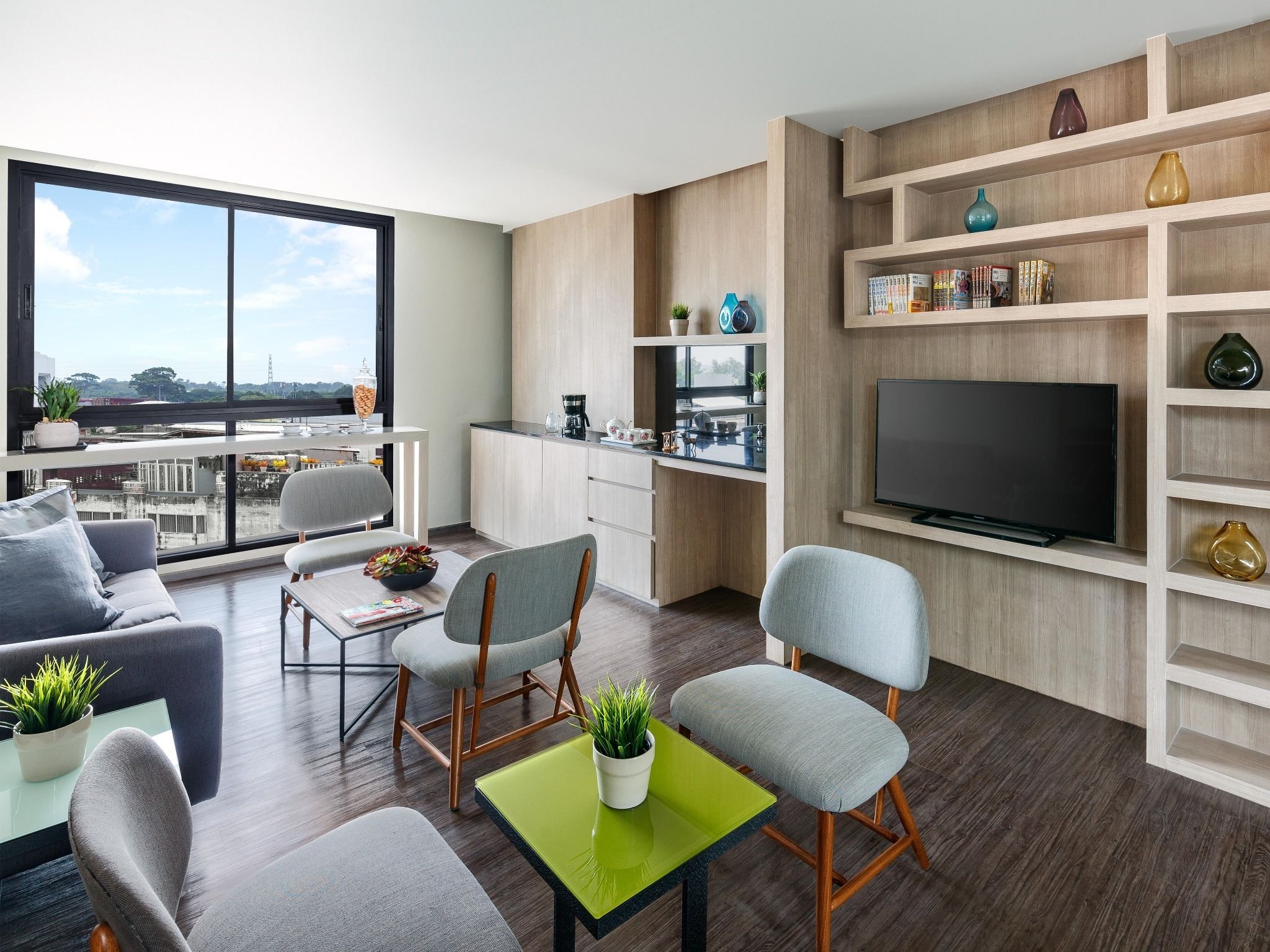 A modern living room with a sofa, chairs, a coffee table, and a TV. There's a balcony with a view of the city.