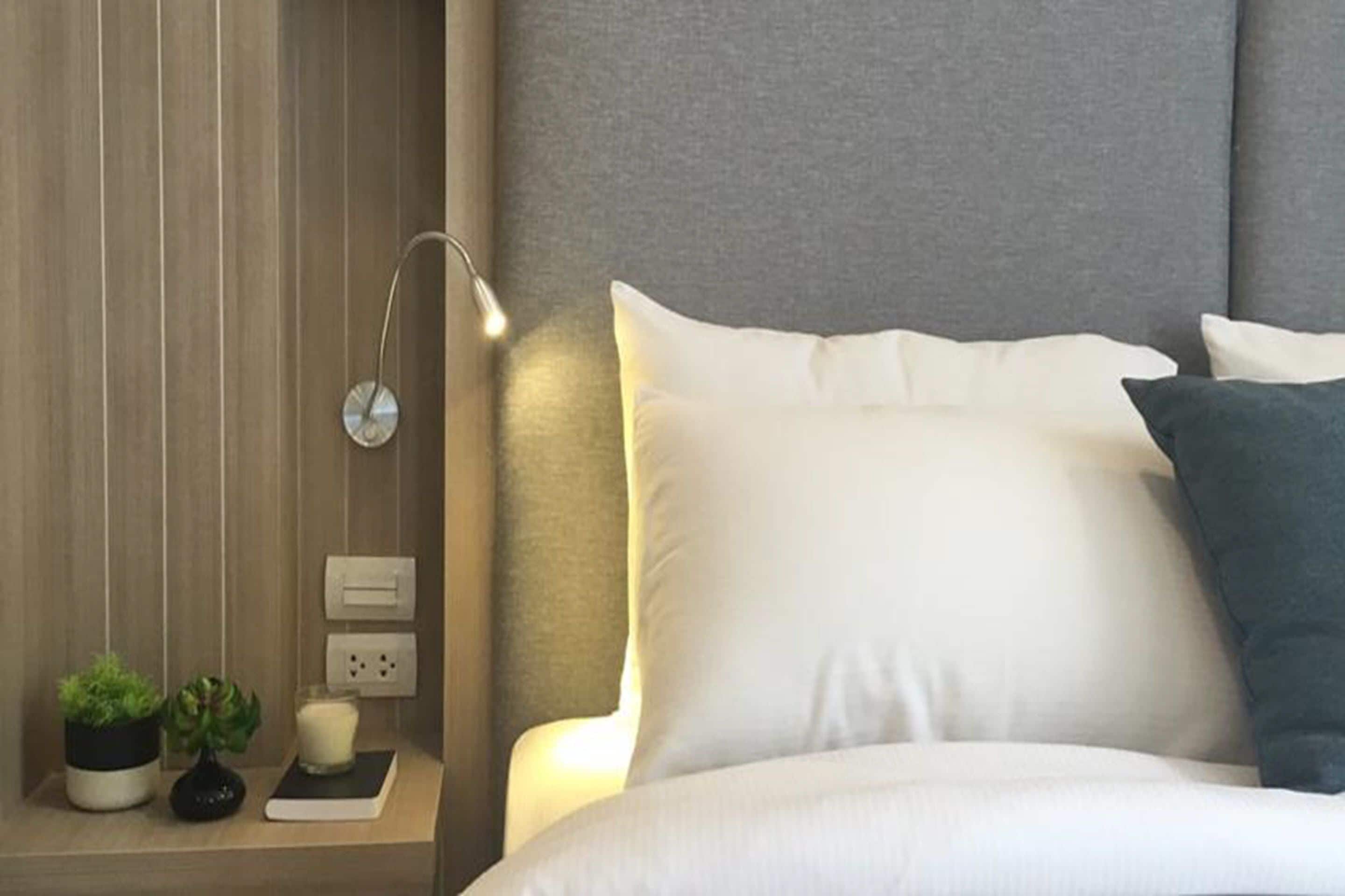 A neatly made bed with white and dark blue pillows, a bedside lamp, and a small bedside table with a plant, a candle, and a book.