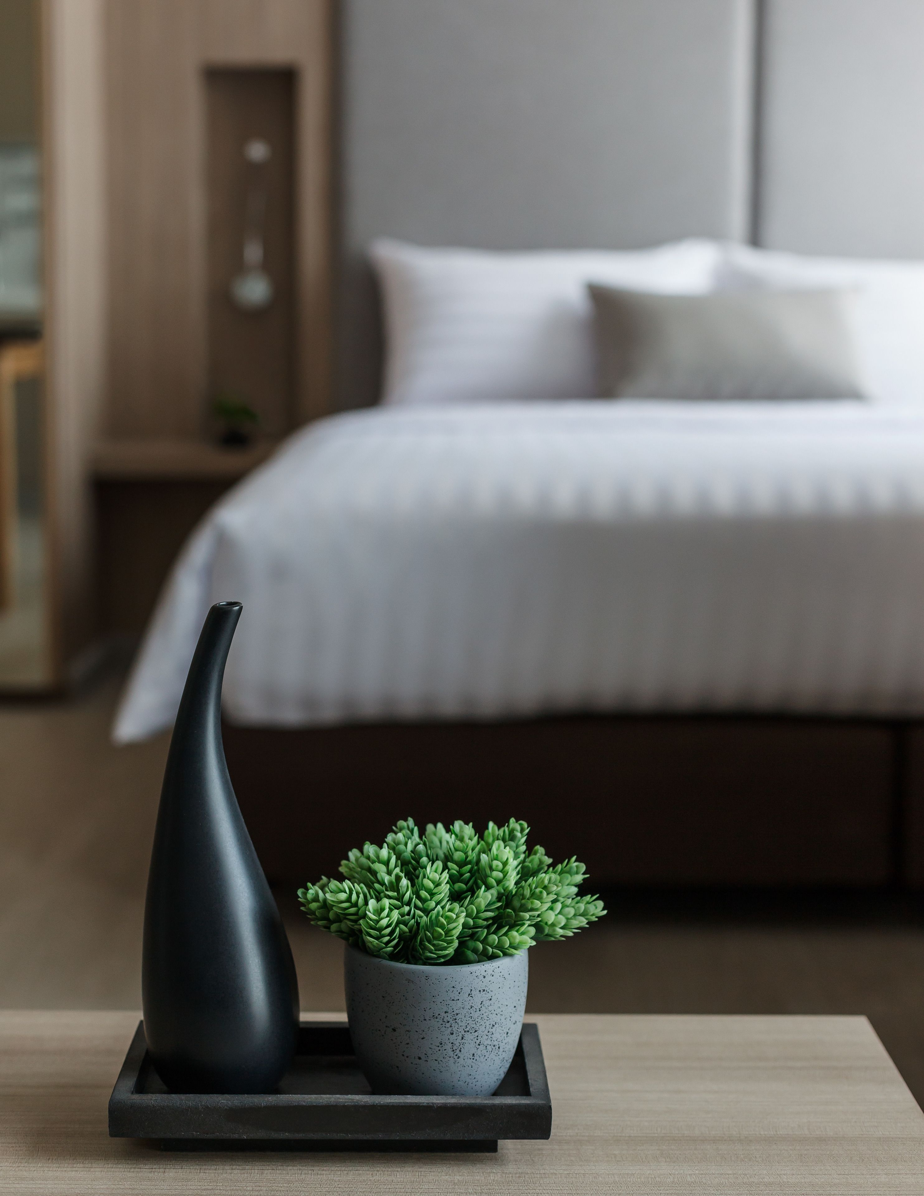 A modern bedroom with a neatly made bed, a small potted plant on a tray, and a black vase on the nightstand.