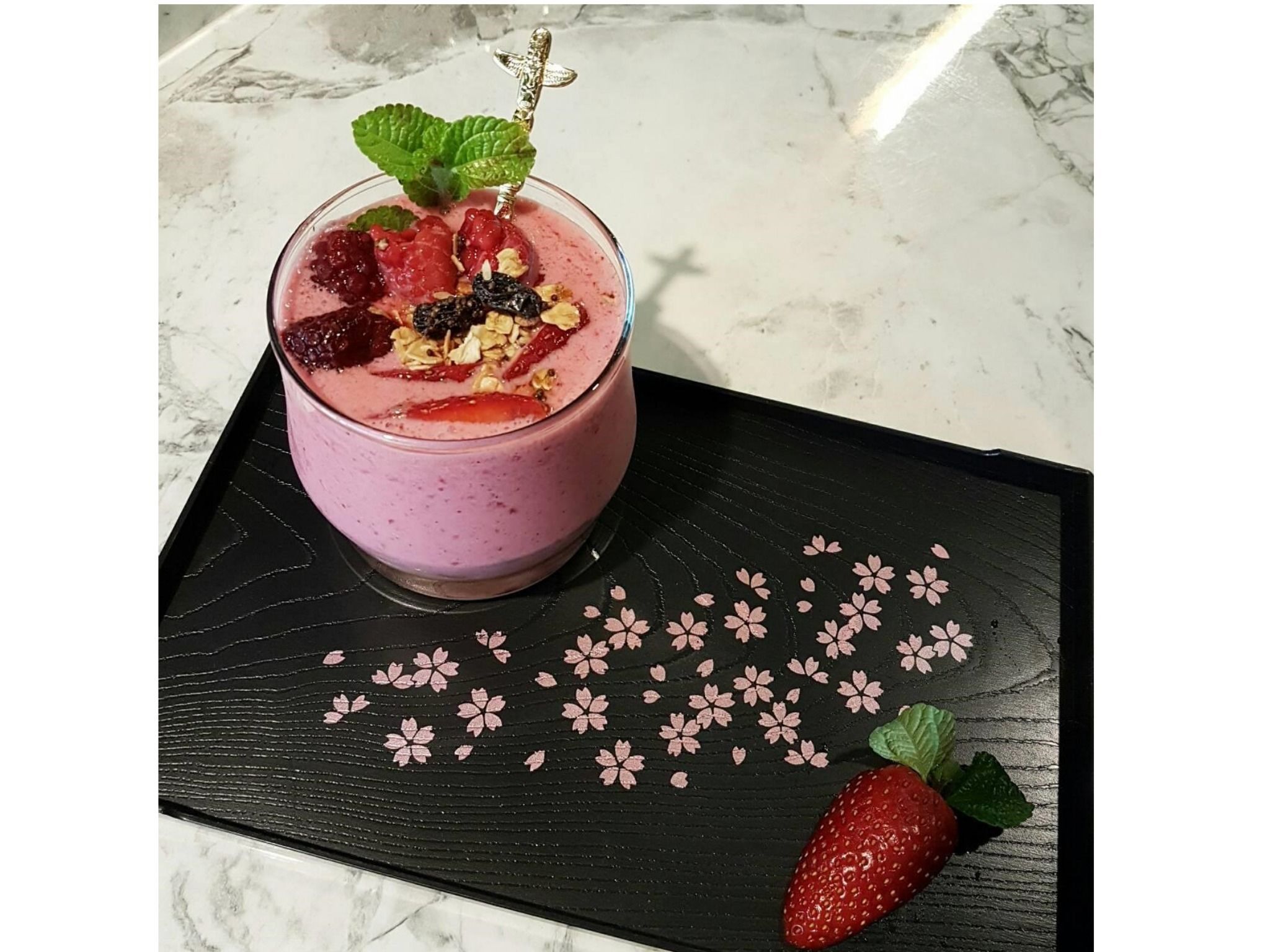 Un bicchiere di smoothie con fragole e menta su un vassoio nero decorato con fiori di ciliegio.