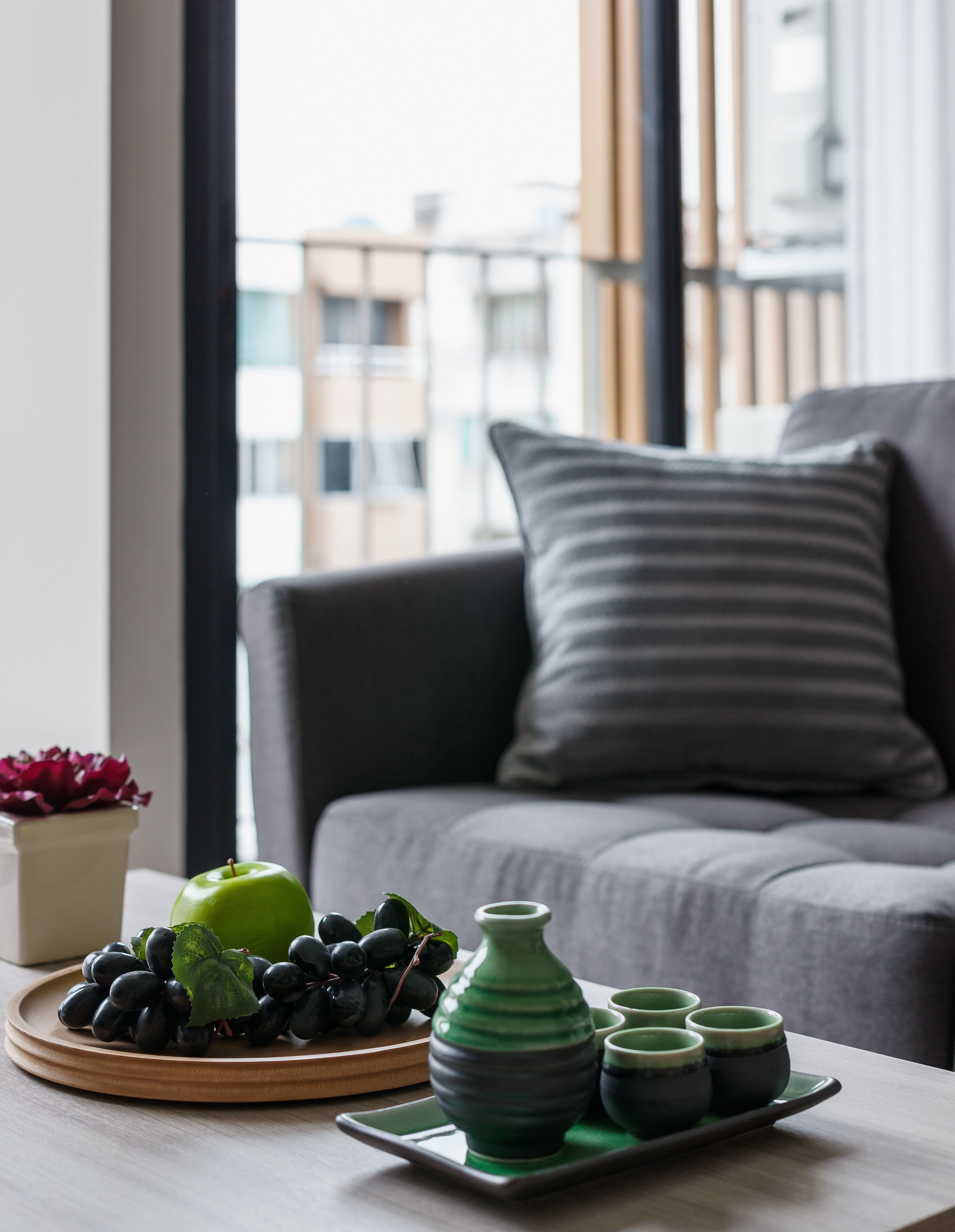 Een moderne woonkamer met een grijze bank, een groene appel, zwarte druiven en een set groene en grijze kommen op een houten tafel.