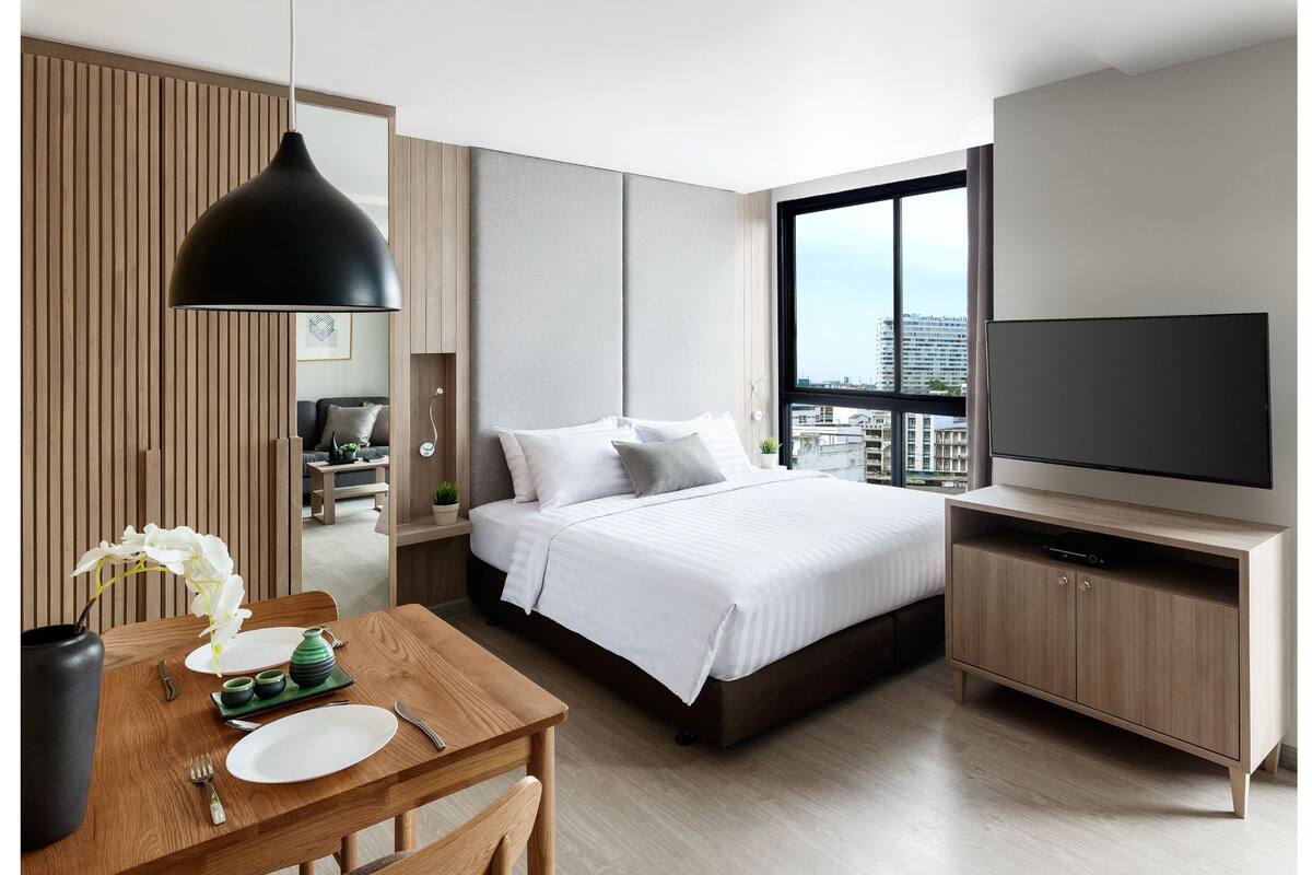Ein modernes Hotelzimmer mit einem großen Bett, einem Fernseher, einer Essecke mit Tisch und Stühlen sowie Blick auf die Stadt durch das Fenster.