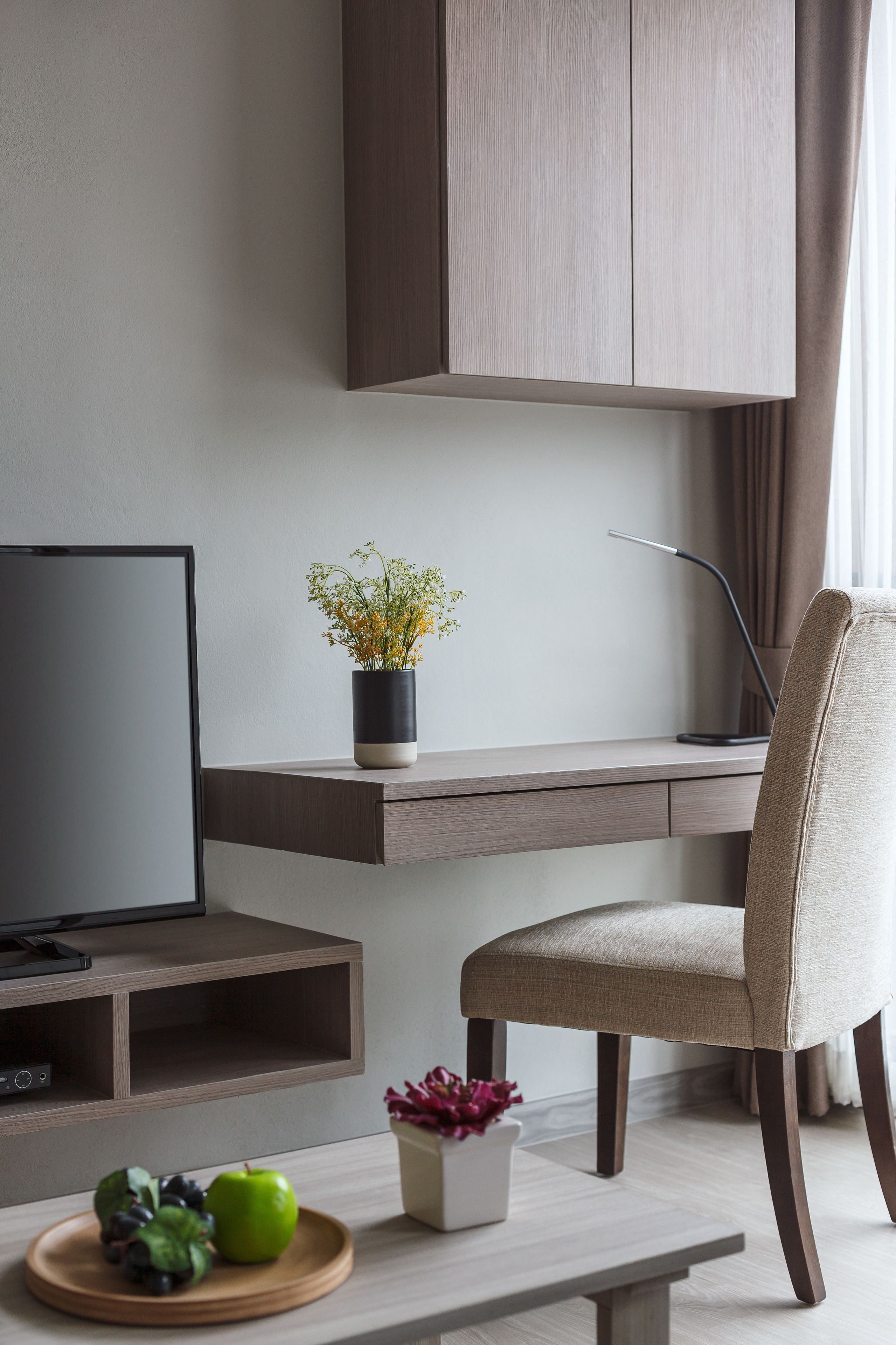 A modern living room with a flat-screen TV, a desk with a laptop, a chair, and a small table with fruits and flowers.