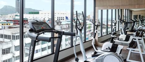 Una palestra con diverse attrezzature per esercizi, ampie finestre e vista su edifici e montagne.