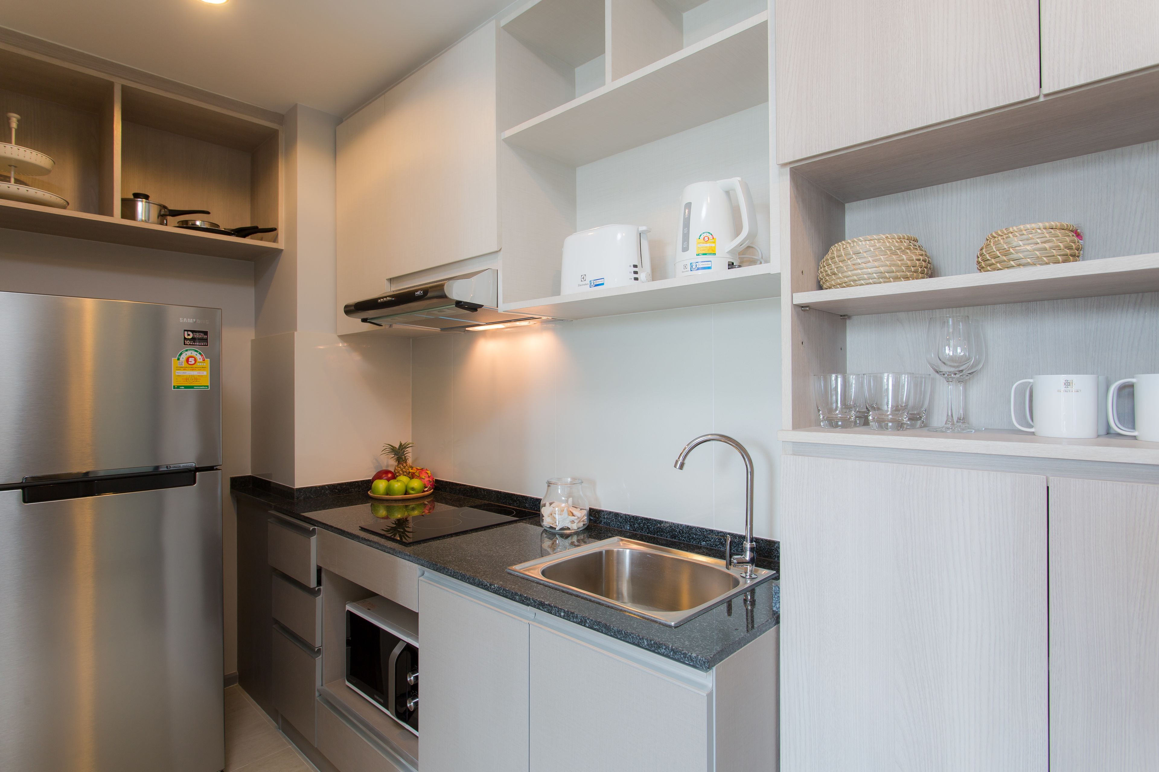 Een moderne keuken met een roestvrijstalen koelkast, een ingebouwde oven, een spoelbak en een aanrecht met een zwarte granieten afwerking. Er zijn witte kasten en een plank met keukengerei.