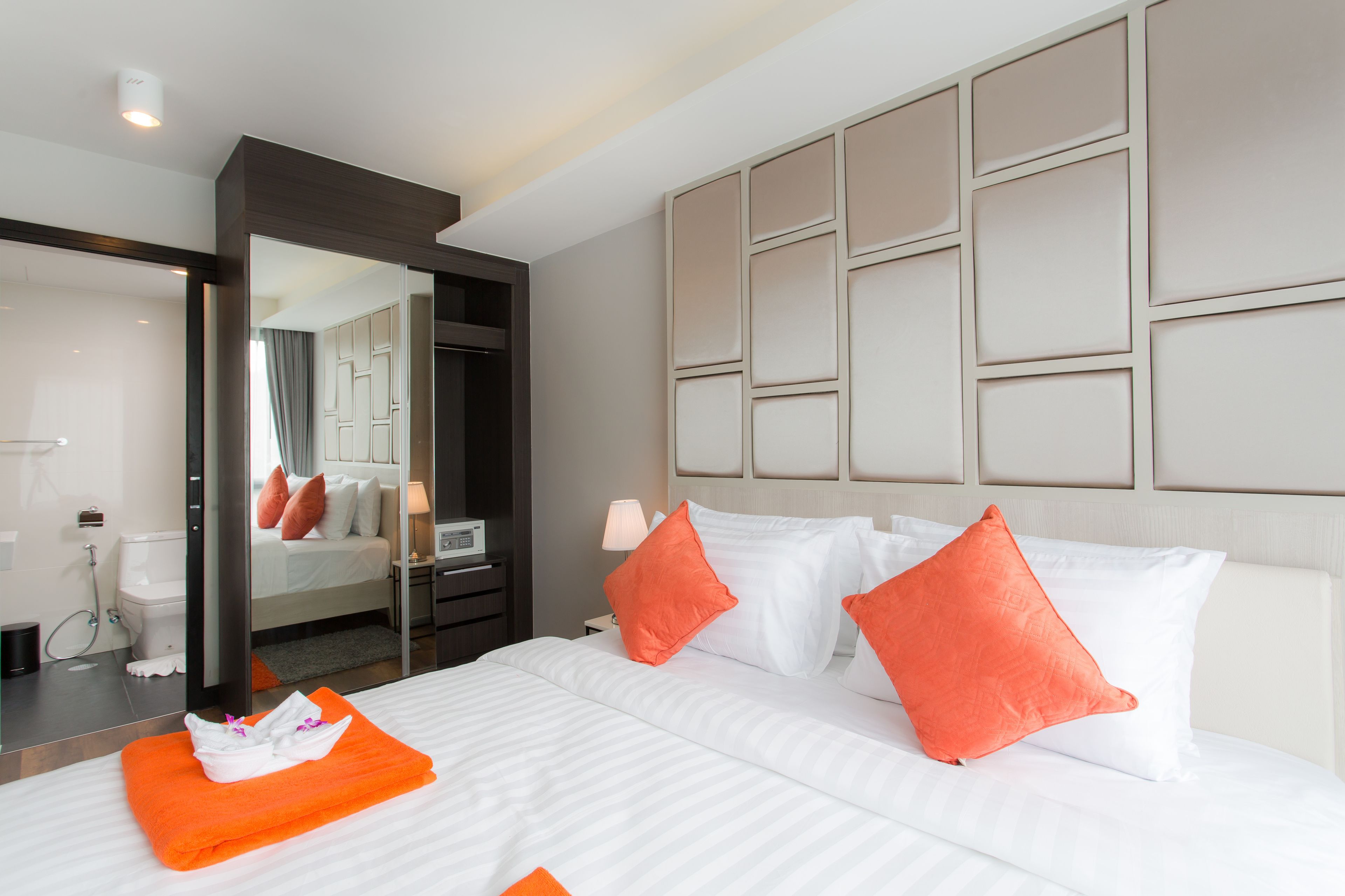 Habitación de hotel con una cama grande, dos almohadas naranjas, un cabecero blanco y un espejo que refleja la habitación.