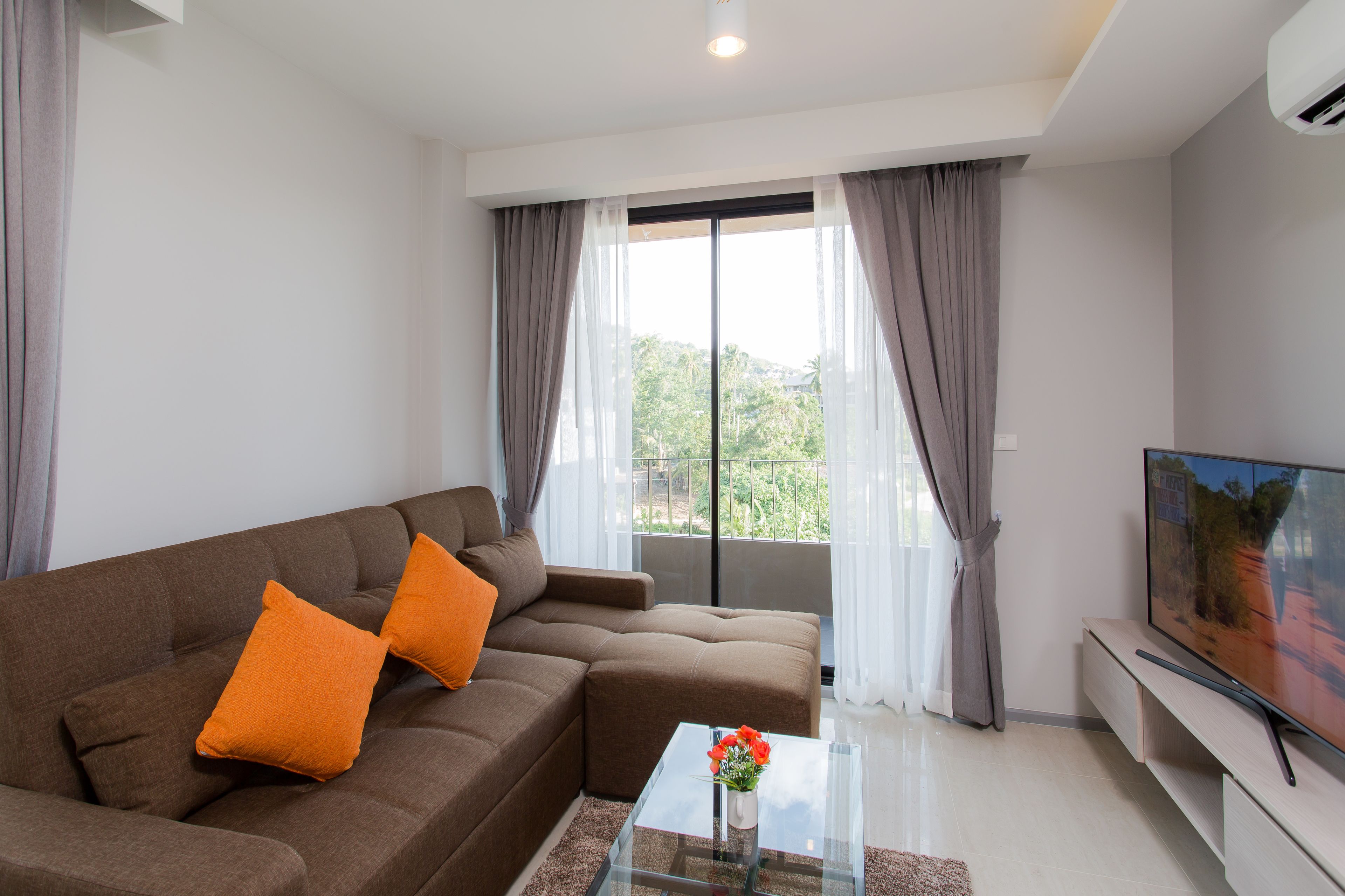 A modern living room with a brown sofa, orange cushions, a glass coffee table, and a flat-screen TV.