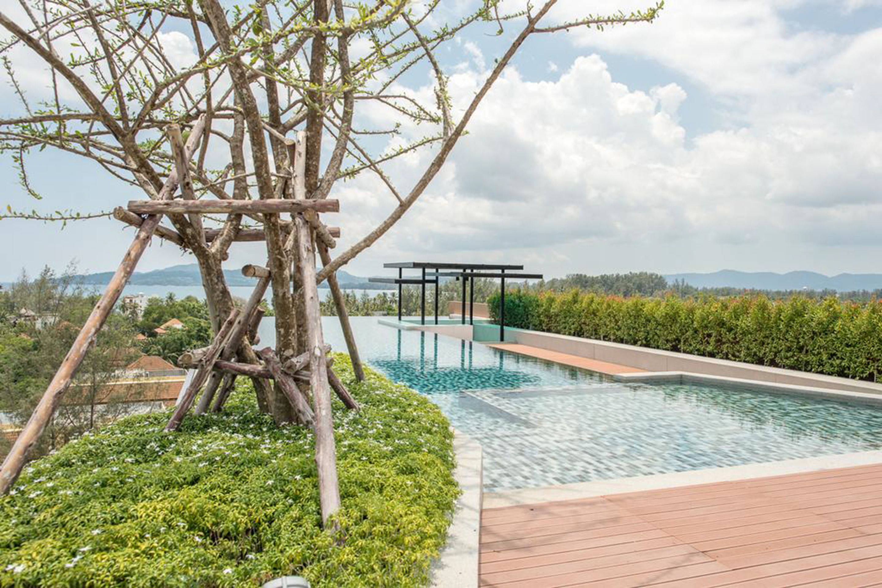 Ein Schwimmbad mit Holzdeck und Pergola, umgeben von Grünpflanzen und Blick auf ferne Berge.