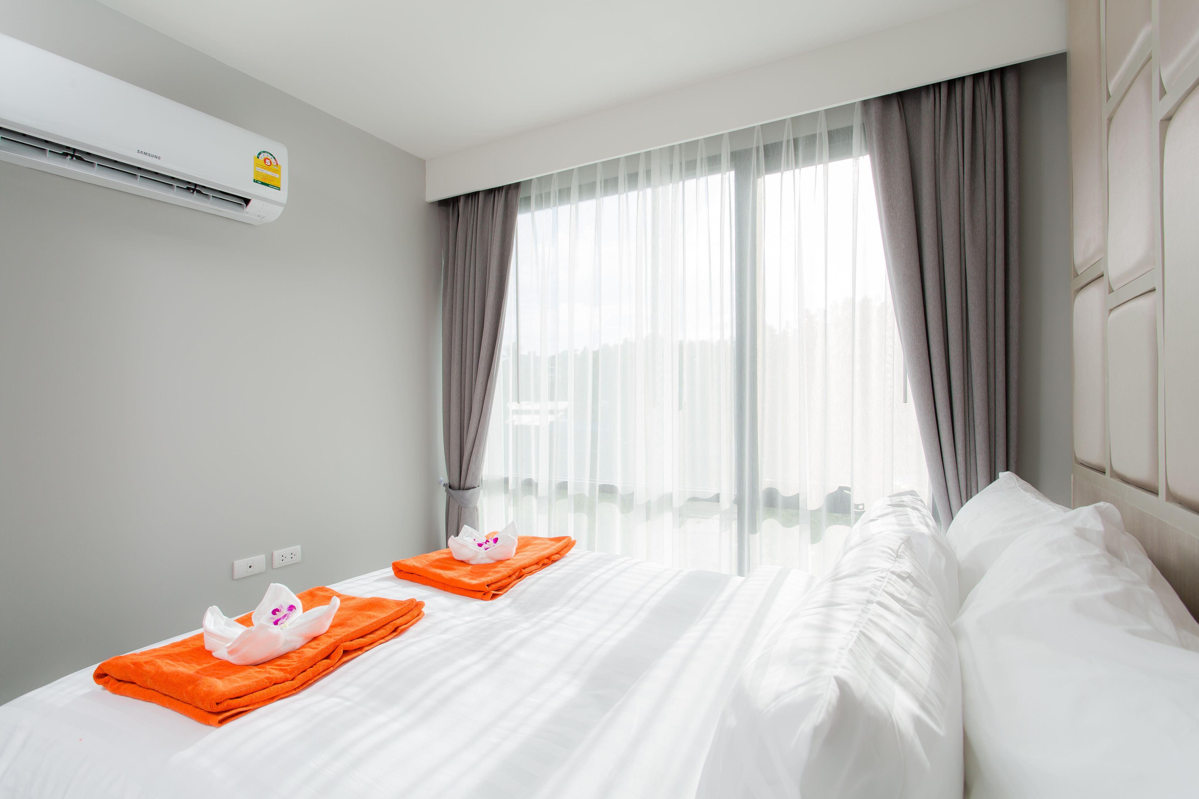 Hotellihuoneessa on suuri sänky, kaksi tyynyä, valkoinen pyyhe ja pieni oranssi pyyhe sängyllä. Huoneessa on ikkuna, jossa on läpikuultavat verhot, ja ikkunasta on näkymä ulos.