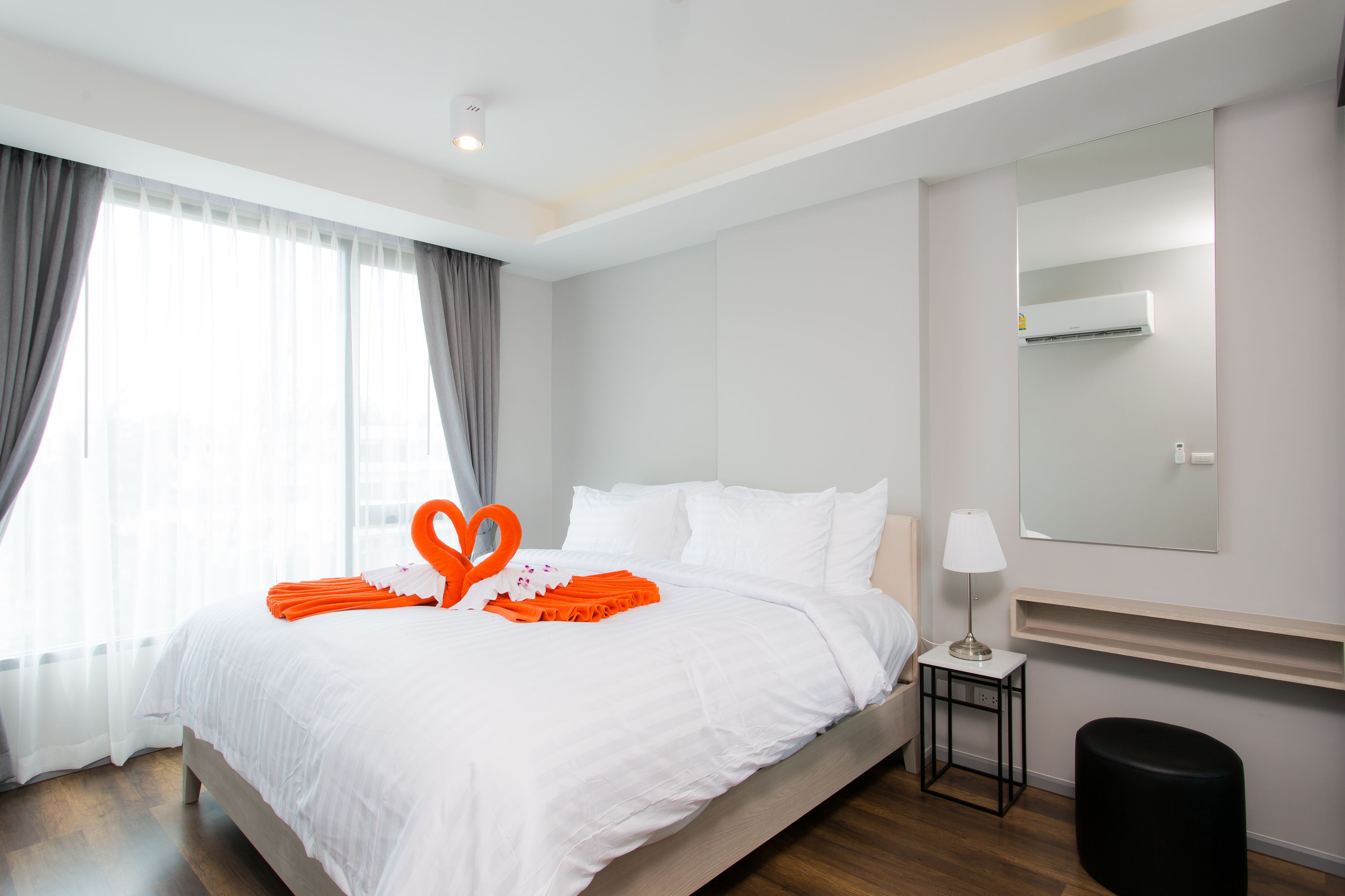 A hotel room with a large bed, a bedside table with a lamp, a mirror on the wall, and a window with curtains.