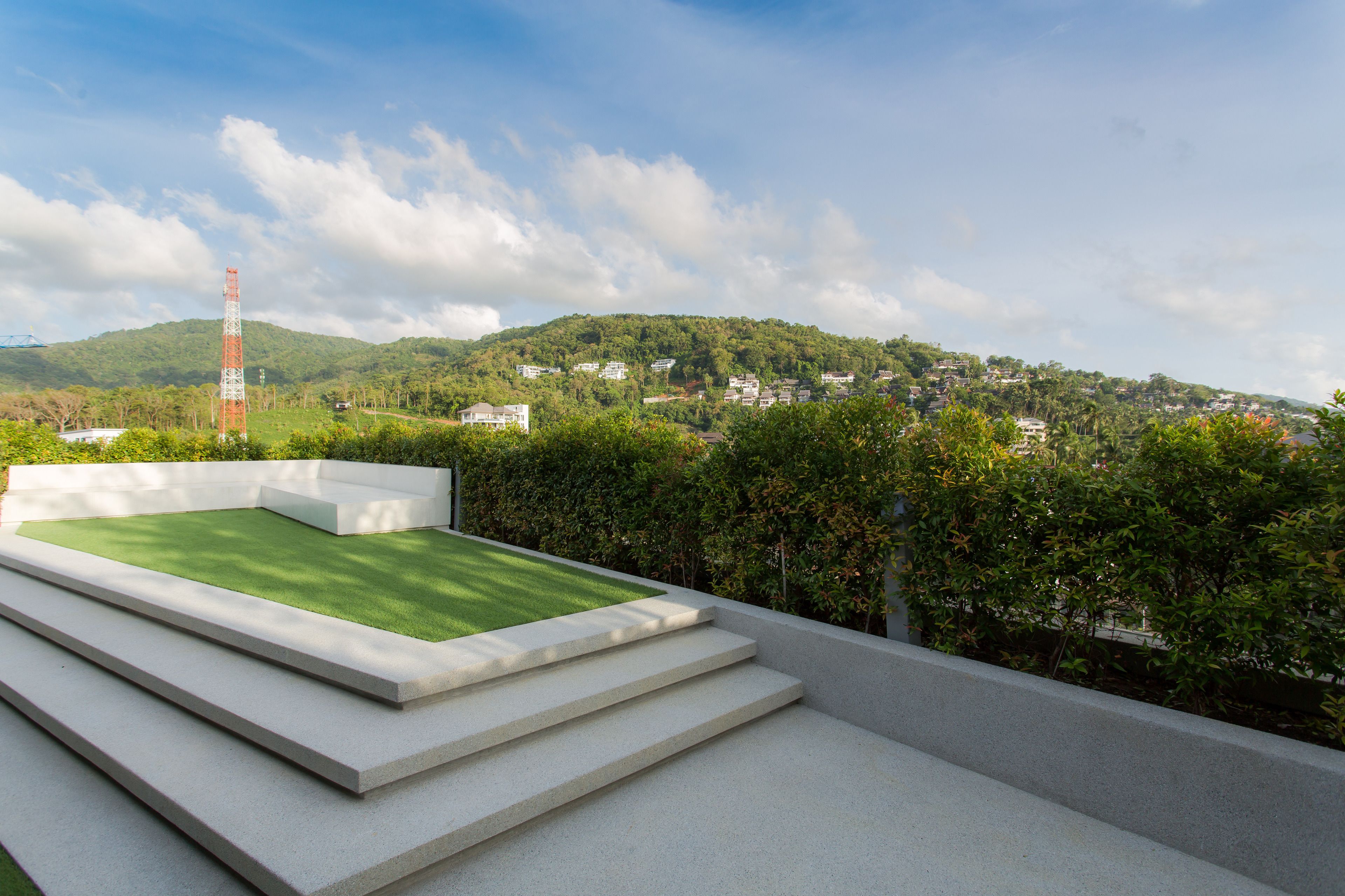 A rooftop terrace with a green lawn and steps leading down, overlooking a hillside with houses and a tall communication tower.