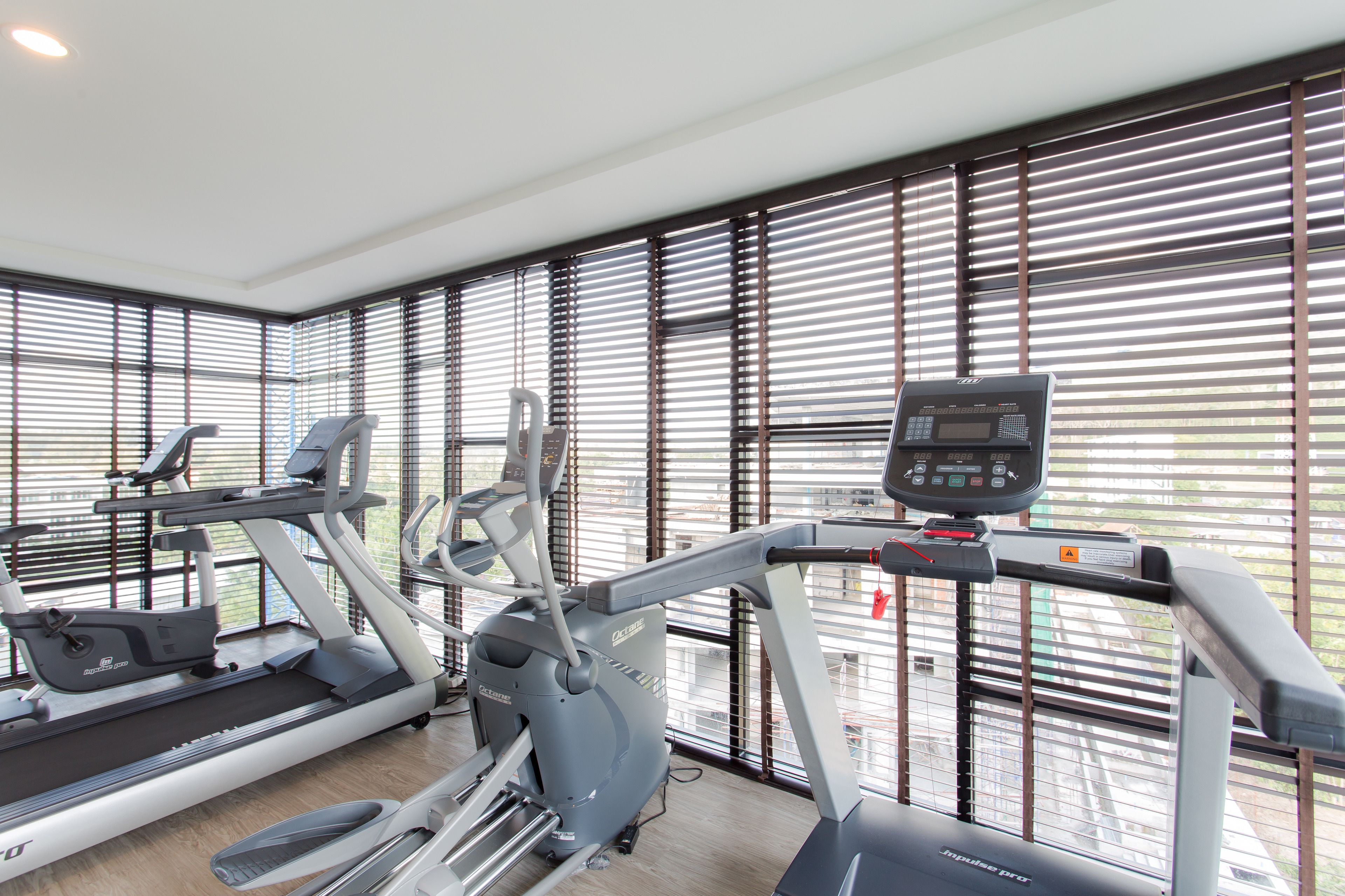 A gym with treadmills and elliptical machines, large windows with blinds, and a control panel on one of the machines.