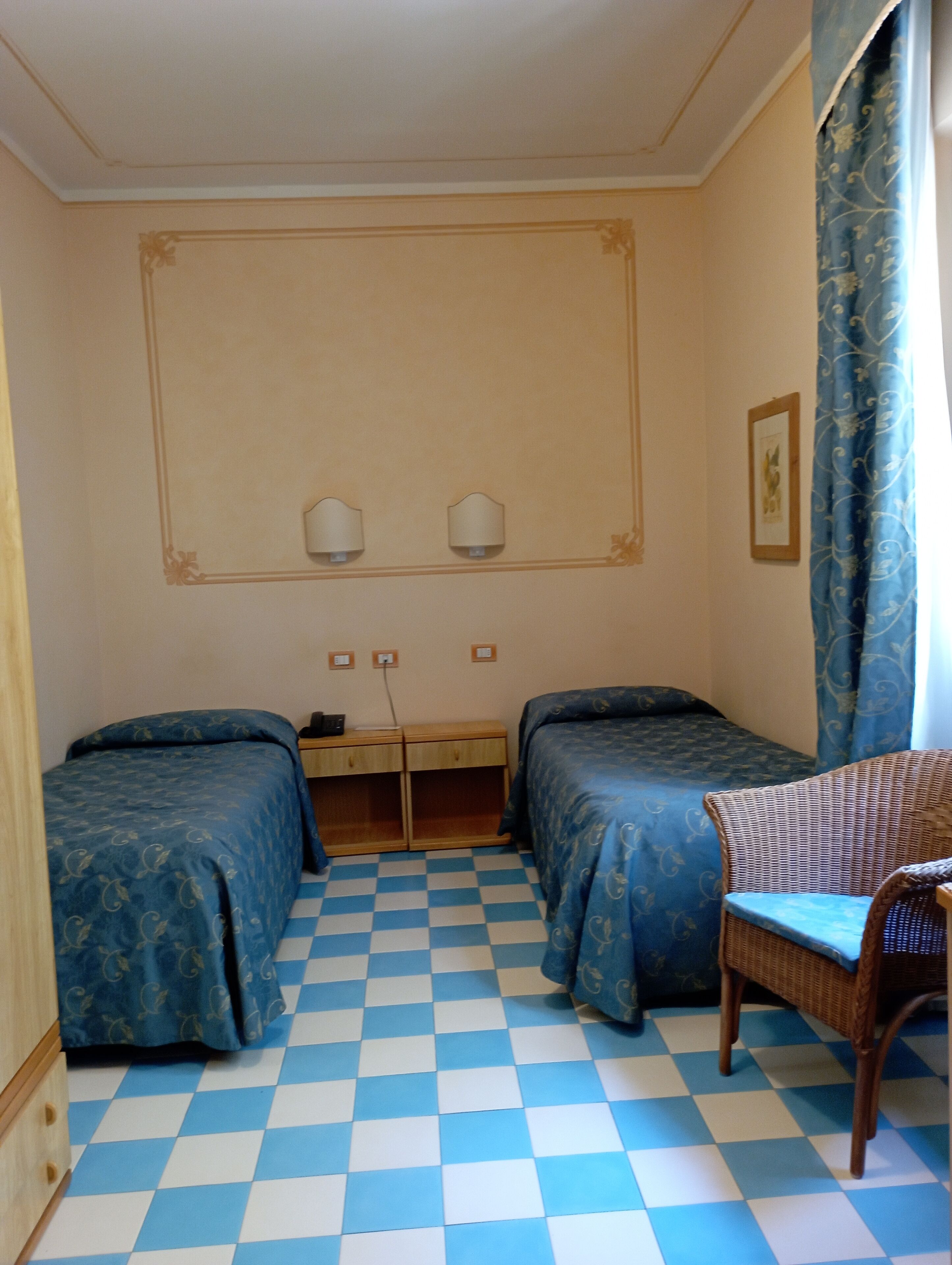 A room with two beds, a desk with two wall lamps, a wicker chair, and a checkered floor.