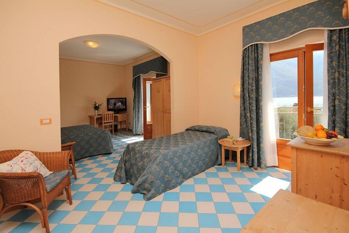 A hotel room with a checkered floor, two single beds, a small table with a fruit bowl, a TV, and a balcony with curtains.