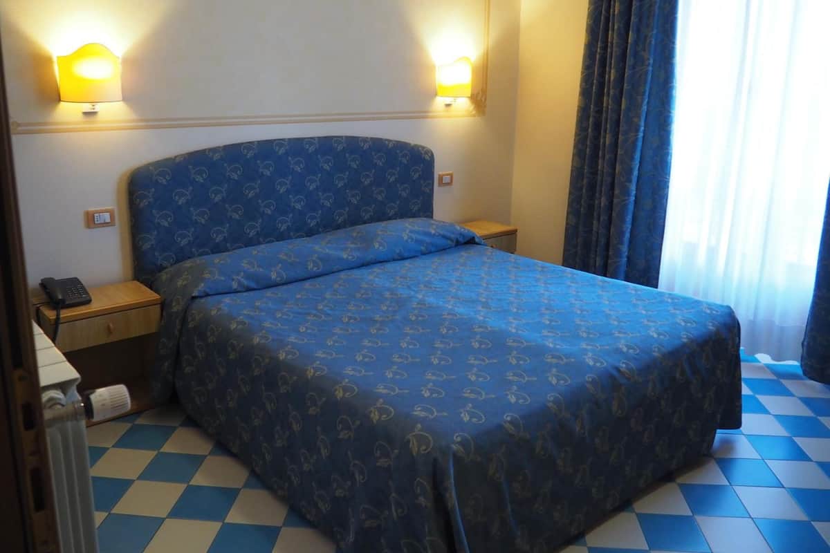 A hotel room with a bed, two bedside lamps, a telephone, a radiator, and a checkered floor.