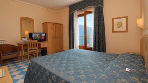 A hotel room with a bed, a desk with a television, a chair, a wardrobe, and a window with a view. - Hotel Locanda Ruscello Garnì (Limone sul Garda)