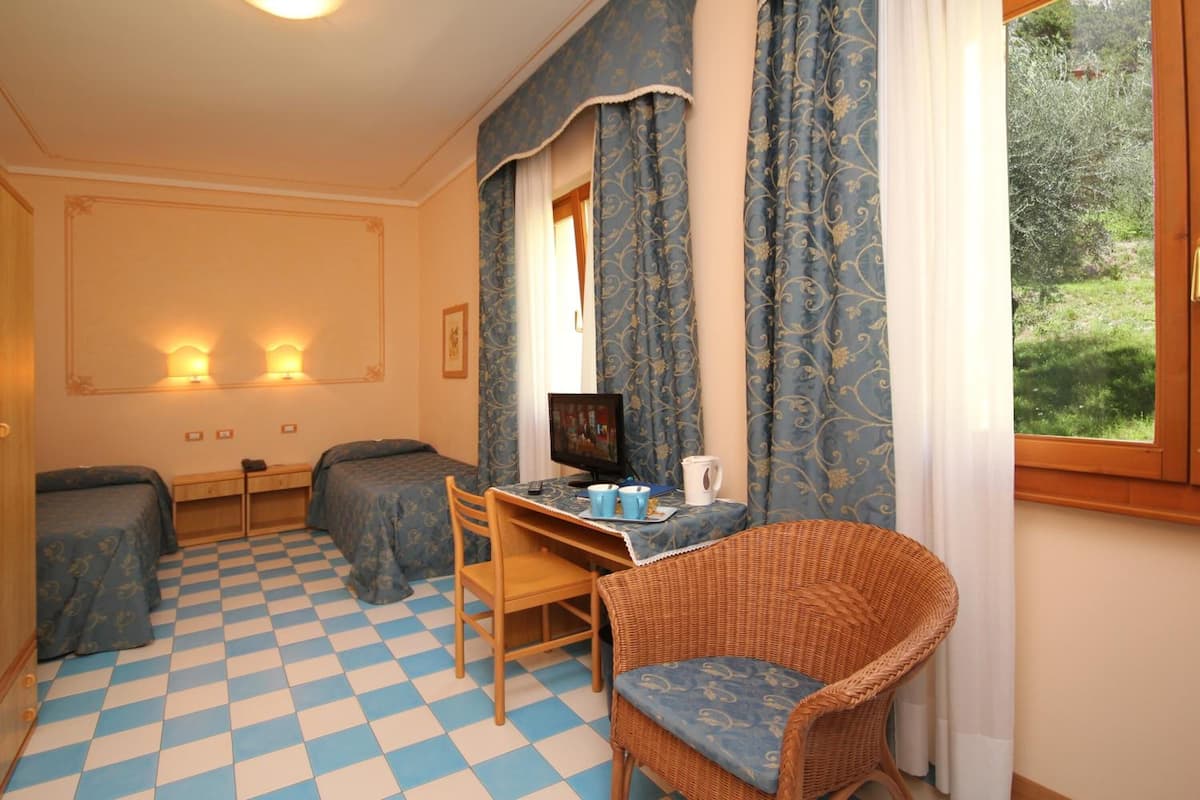 A room with two beds, a desk with a computer, a wicker chair, and a window with curtains.