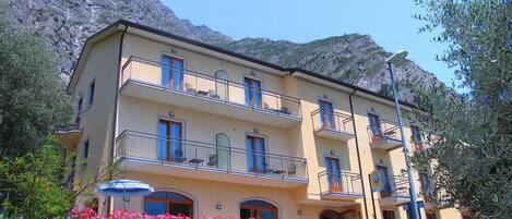 A multi-story hotel with balconies, stone walls, and pink flowers in front.